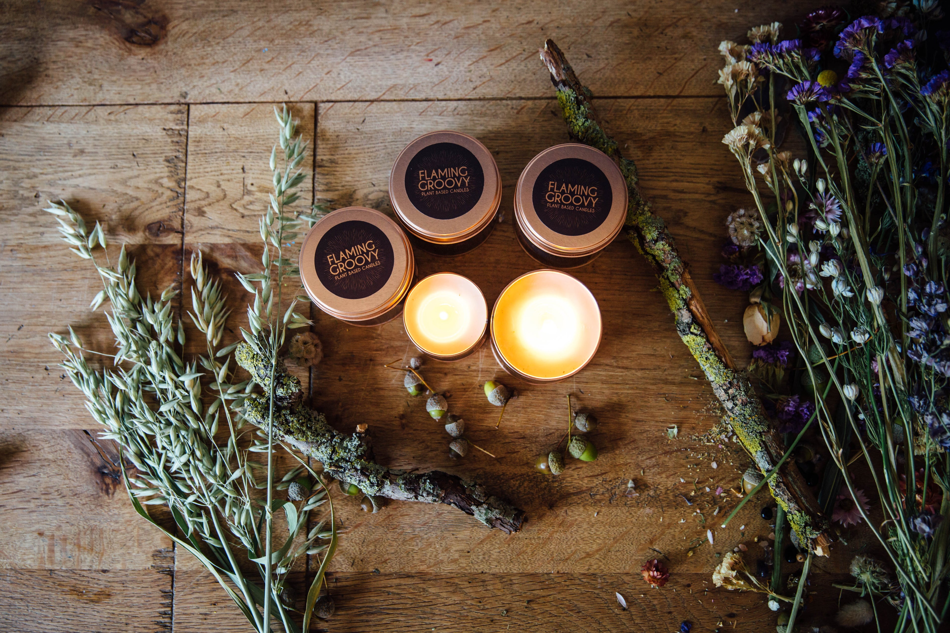 Rose gold flaming groovy candle tins, some lit, some not. Herbs and grasses on wooden background.