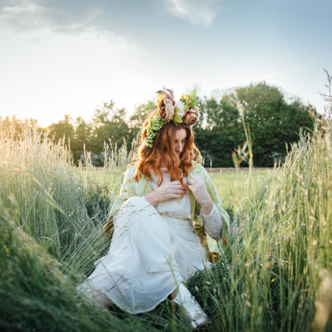 Carolyn is sitting in a meadow in long grass. It’s golden hour, the sun is starting to set. She wears a Tatty La La flower crown with vintage floor length cream dress, beaded cape and gold boots. The crown is made with up-cycled flowers. 