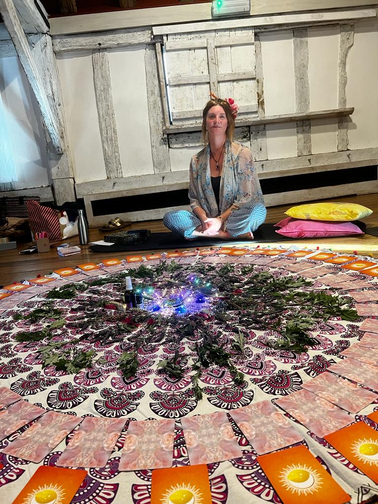 Carolyn is sitting on the floor in a sacred circle with a mandala of herbs and oracle cards.